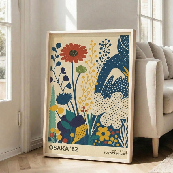 May include: Framed vintage-style poster featuring colorful botanical illustration with stylized flowers, leaves, and plants in navy blue, orange, yellow, and green on cream background. Text reads "Osaka '82" and "Flower Market" in Japanese and English. Light wood frame leaning against white wall next to beige sofa in bright, minimalist living room with large windows.