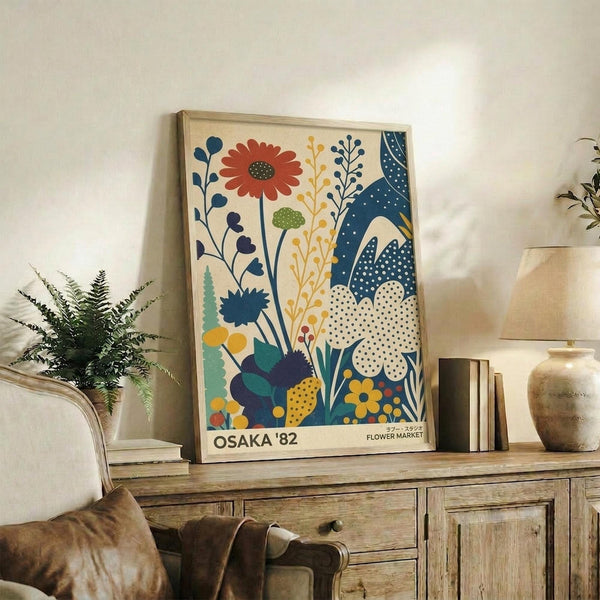 May include: A framed botanical poster with colorful stylized flowers in blues, oranges, and yellows displayed on a rustic wooden sideboard next to books, a ceramic vase, and a potted fern plant in a cozy living room setting with neutral walls and a beige armchair.