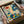 May include: Hands unwrapping a colorful botanical art print from shipping materials and cardboard packaging. The artwork features stylized flowers, leaves, and plants in vibrant blues, oranges, yellows, and greens with dotted patterns and folk art styling. The print is matted in cream and set in a black frame, photographed on white fabric with part of a glass object visible in the background.