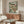 May include: Framed botanical fish print displayed above wooden credenza in modern living room. Artwork features three speckled fish with large eyes swimming among green foliage and coral dots on light background. Scene includes white curved chair, glass-top oval coffee table, and decorative lighting fixtures. Natural sunlight creates shadows across warm wood furniture and neutral walls.