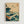 May include: Framed Japanese-inspired wave artwork hanging on white wall. Blue-green stylized ocean waves with white foam crash dramatically across beige background. Three small birds fly in upper portion. Red square cartouche in top right corner. Natural wood frame. Art style mimics traditional woodblock prints with flowing curved lines and organic wave patterns.