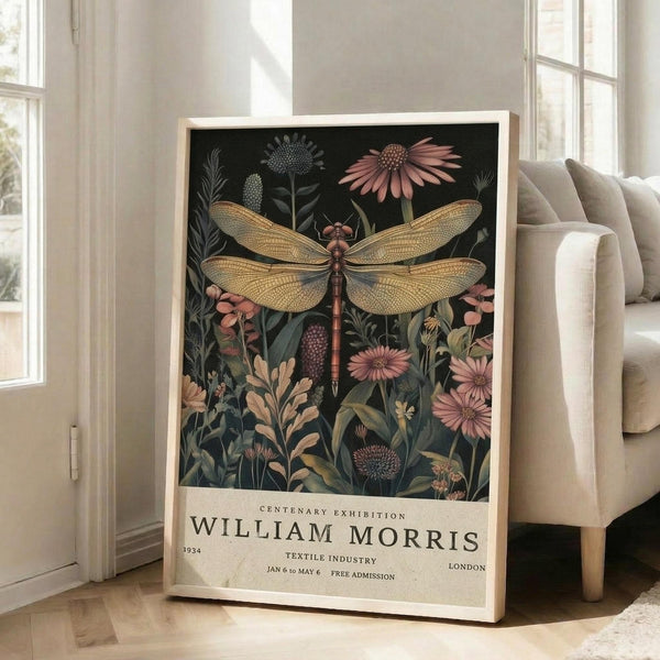May include: Framed botanical poster featuring a large golden dragonfly with detailed wings surrounded by pink flowers, green foliage, and plants against a dark background. Exhibition text reads "William Morris 1934" at bottom. Light wood frame leans against white wall next to beige upholstered sofa in bright, modern living room with tall windows and natural lighting.