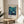 May include: Framed vintage Japanese exhibition poster featuring underwater scene with whales, coral, and sea life in blue and teal tones, displayed on round wooden side table next to beige upholstered chair and plant in bright, minimalist room with white walls and natural lighting.
