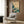 May include: Japanese-inspired wave artwork with teal and turquoise waves, white foam caps, and flying birds against a cream background, displayed above a beige upholstered armchair with geometric patterned pillow. Scene shows modern living room with brass floor lamp, dark round side table with open book, wood panel wall, and natural lighting from window with sheer curtains.