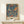 May include: Framed William Morris exhibition poster featuring two birds perched on branches surrounded by red strawberries, green leaves, and small flowers on a deep blue background. The vintage-style poster sits on a light wooden shelf next to a small white wrapped gift with greenery, displayed against a cream wall in natural lighting.
