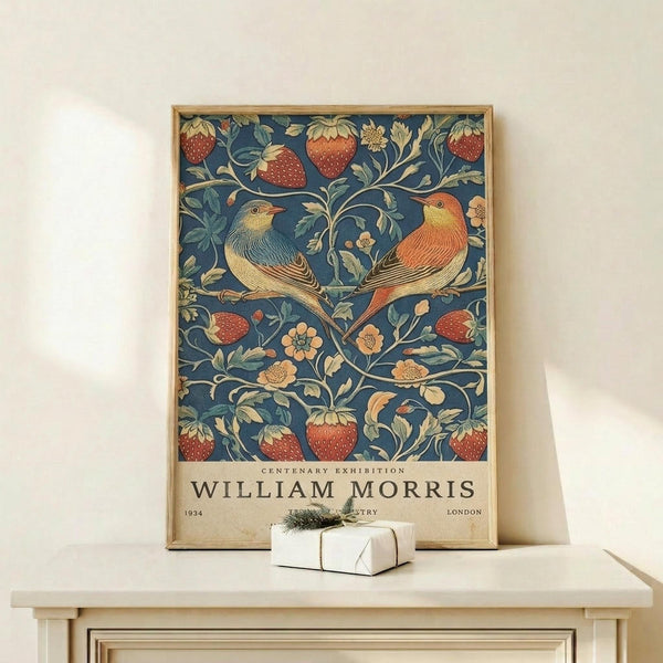 May include: Framed William Morris exhibition poster featuring two birds perched on branches surrounded by red strawberries, green leaves, and small flowers on a deep blue background. The vintage-style poster sits on a light wooden shelf next to a small white wrapped gift with greenery, displayed against a cream wall in natural lighting.