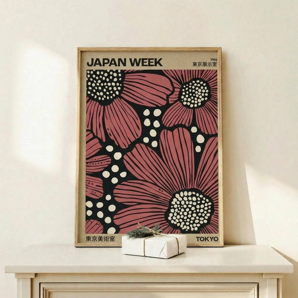 May include: Framed vintage-style poster with bold pink sunflower-like blooms and white polka dots on black background, displayed on light wood console table next to small wrapped gift box with evergreen sprig. The poster features "JAPAN WEEK" text and Japanese characters, housed in natural wood frame against white wall.