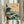May include: Person in beige ruffled sleeves holding framed Japanese-style wave artwork in light wood frame. Teal and navy blue stylized ocean waves with white foam crash against brown rocky coastline. Flying birds silhouetted against cream sky with red Asian text seal. Small boat visible in distance. Dried pampas grass stems beside frame against soft white curtained window background.