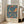 May include: Framed poster with blue background featuring two colorful birds perched among strawberries, flowers and decorative foliage in William Morris textile style. Exhibition text below reads "Centenary Exhibition William Morris 1934 Textile Industry London." Light wood frame leans against white wall next to beige upholstered chair in bright, minimalist living room with large windows.