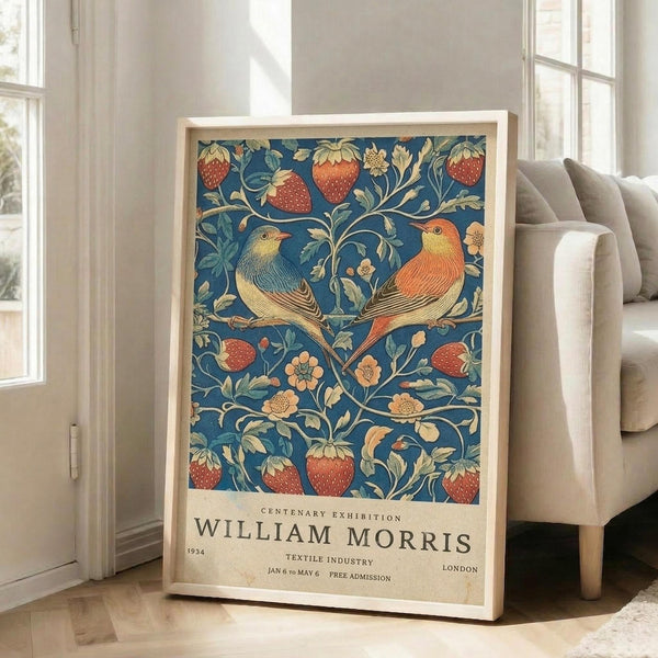 May include: Framed poster with blue background featuring two colorful birds perched among strawberries, flowers and decorative foliage in William Morris textile style. Exhibition text below reads "Centenary Exhibition William Morris 1934 Textile Industry London." Light wood frame leans against white wall next to beige upholstered chair in bright, minimalist living room with large windows.