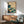 May include: Framed Japanese-style wave artwork hanging above a wooden sideboard with table lamp, next to a mid-century modern chair in sage green upholstery. The print features dramatic teal and turquoise ocean waves with white foam, flying birds, and a red text cartouche against cream background. Natural sunlight creates geometric shadows on white wall.