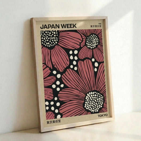 Vintage-style poster featuring large pink flowers with radiating petals and white dotted centers on a black background with scattered white polka dots. The design shows "JAPAN WEEK" at the top with "1984" and Japanese characters in the upper right, and "TOKYO" with more Japanese text at the bottom. The poster is displayed in a light wooden frame leaning against a white wall, showing the full artwork in a minimalist setting.