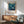 May include: Framed vintage-style poster featuring underwater ocean scene with whales, coral, and marine life in blue and teal tones, displayed above wooden sideboard with small white table lamp, next to sage green mid-century modern armchair with wooden frame in sunlit room with white walls.