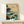 May include: Japanese-style wave artwork in wooden frame displayed on light wood console table against white wall. Print features dramatic teal and dark blue ocean waves with white foam, flying birds, distant mountains, and traditional red seal. Small white wrapped gift box sits beside the framed print. Art shows dynamic water movement with traditional woodblock print aesthetic.
