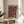 May include: Framed vintage-style poster with pink and black floral design featuring stylized flowers with white polka dot centers, displayed on a round wooden side table next to a beige upholstered chair. Warm natural lighting creates soft shadows on cream-colored wall, with green plant leaves visible in corner of minimalist modern room setting.