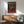 May include: Framed poster featuring a Rottweiler dog wearing a chunky red knit sweater against a dark background, with "DOGUE" text at top in cream letters. The wooden-framed artwork hangs above a mid-century modern walnut credenza with a white ceramic table lamp. A teal upholstered armchair with wooden frame sits nearby on a wooden floor, with natural sunlight casting shadows across white walls.