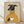 May include: Framed poster print leaning against white wall on light wood floor. Shows illustration of black greyhound dog wearing mustard yellow ribbed turtleneck sweater, holding white coffee cup with steam rising. Dog has alert ears and bright eyes against cream textured background. Natural wood frame, soft shadows from window light.