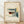 May include: Framed poster print featuring modernist architectural illustration in muted tones of cream, teal, yellow and rust red. Shows geometric building with grid windows, curved staircase, and angular forms in Bauhaus style. Natural wood frame sits on light wooden floor against white textured wall with soft shadows.