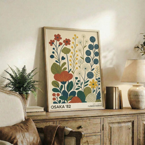 May include: Framed botanical poster with stylized flowers in coral, teal, yellow and green displayed on rustic wooden sideboard next to fern plant, ceramic vases, and books in neutral-toned living room setting with beige armchair