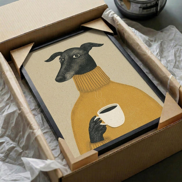 May include: Framed art print of a greyhound wearing a mustard yellow turtleneck sweater, holding a white coffee cup with steam rising. The dog has dark fur and alert ears against a cream background. The print sits in a black frame inside a cardboard shipping box with protective tissue paper, surrounded by wooden frame corners and packaging materials on a table surface.