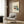 May include: Framed Bauhaus architectural poster displayed on wood-paneled wall above cream upholstered chair with geometric pillow. Poster features modernist building illustration in muted greens, yellows, and reds with stairs and glass windows. Golden floor lamp and dark round side table with open book complete the mid-century modern room setting with natural lighting from window.