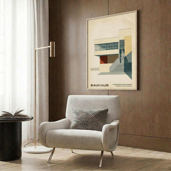 May include: Framed Bauhaus architectural poster displayed on wood-paneled wall above cream upholstered chair with geometric pillow. Poster features modernist building illustration in muted greens, yellows, and reds with stairs and glass windows. Golden floor lamp and dark round side table with open book complete the mid-century modern room setting with natural lighting from window.