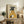 May include: Framed artwork showing a black greyhound wearing a mustard yellow ribbed turtleneck sweater, holding a white coffee cup, displayed on a rustic wooden sideboard alongside books, a small potted fern, ceramic vases, and brown leather furniture in a warm, neutral-toned living room setting.