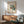 May include: Modernist architectural poster in wood frame showing geometric white building with stairs, teal windows, and red accent, displayed above walnut credenza with ceramic table lamp in bright room with mid-century green upholstered chair and round coffee table.