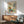 May include: Framed botanical art print featuring stylized flowers and plants in earthy tones of rust, teal, yellow and green on cream background, displayed above mid-century modern wood credenza with table lamp, next to sage green upholstered armchair in bright sunlit room with white walls