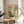 May include: Vintage-style botanical poster with colorful stylized flowers and plants in cream, orange, teal, and green, displayed in wooden frame on round wooden side table next to beige upholstered chair with decorative pillow, plant leaves visible in corner, soft natural lighting creating shadows on neutral wall