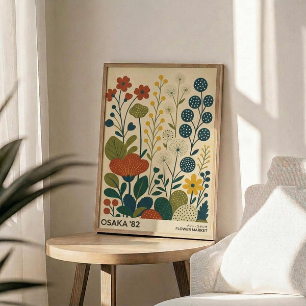 May include: Vintage-style botanical poster with colorful stylized flowers and plants in cream, orange, teal, and green, displayed in wooden frame on round wooden side table next to beige upholstered chair with decorative pillow, plant leaves visible in corner, soft natural lighting creating shadows on neutral wall