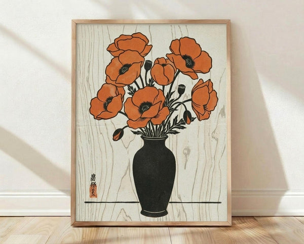 May include: A framed botanical print displayed against a white wall on light wooden flooring. The artwork features orange poppies with black centers arranged in a dark green vase, rendered in vintage Japanese woodblock print style with visible wood grain texture in the background. The piece is housed in a natural wood frame and casts soft shadows on the textured white wall behind it.