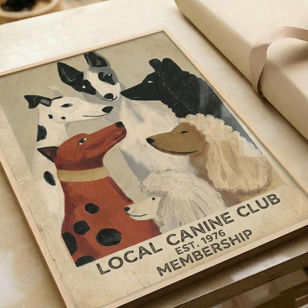 A vintage-style poster featuring six illustrated dog portraits in muted earth tones on cream background. Dogs include dalmatians, terriers, and other breeds rendered in painterly style with black, white, brown and rust colors. Text reads "LOCAL CANINE CLUB EST. 1976 MEMBERSHIP" at bottom. The framed print sits on white wrapping paper with ribbon, showing warm lighting and cozy styling.