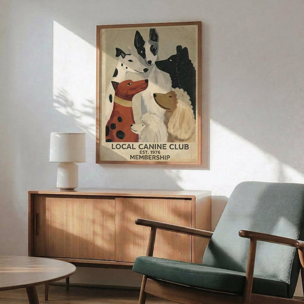 May include: Framed vintage-style poster featuring illustrated dogs in various breeds and colors including spotted, black, white, and reddish-brown dogs arranged in a group portrait style. The artwork has a cream background with text reading "LOCAL CANINE CLUB EST. 1976 MEMBERSHIP" at the bottom. Displayed in a mid-century modern living room with teal upholstered armchair, wooden sideboard, white ceramic table lamp, and natural lighting creating geometric shadows on white walls.