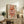 May include: Framed retro-style poster with colorful swirling pasta noodles in red, orange, and green tones against cream background, displaying "PASTA LA VISTA" text with tagline below. Shows wooden sideboard styling with potted fern, ceramic vase, stacked books, and brown leather chair in warm-toned living room setting.