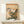 May include: Japanese woodblock print in light wooden frame showing a brown bird perched above orange trumpet vine flowers climbing a traditional thatched roof house, with misty mountains and clouds in background, displayed on cream wooden console table beside small white wrapped gift box against white wall