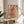 May include: Framed poster with cream background featuring swirling pasta noodles in warm orange, red, and green tones around bold white text reading "PASTA LA VISTA" with smaller subtitle text below. The wooden-framed print sits on a round wooden side table next to a beige upholstered chair, with a potted plant visible in soft natural lighting creating gentle shadows on the white wall behind.