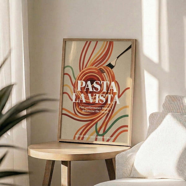 May include: Framed poster with cream background featuring swirling pasta noodles in warm orange, red, and green tones around bold white text reading "PASTA LA VISTA" with smaller subtitle text below. The wooden-framed print sits on a round wooden side table next to a beige upholstered chair, with a potted plant visible in soft natural lighting creating gentle shadows on the white wall behind.