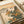 Japanese woodblock print displayed in an open art book showing a brown bird perched among orange trumpet vine flowers with green leaves. Mountains and traditional thatched-roof buildings appear in the background under a peachy sunset sky. The vintage print features delicate linework and muted earth tones typical of traditional ukiyo-e style, with Japanese calligraphy and a red artist seal visible on the cream-colored paper.
