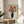May include: Framed vintage-style botanical print featuring orange poppies in black vase displayed on round wooden side table next to beige upholstered chair. Artwork shows Japanese woodblock print aesthetic with orange flowers against light wood grain background. Natural lighting creates soft shadows on neutral wall. Plant leaves visible in corner.