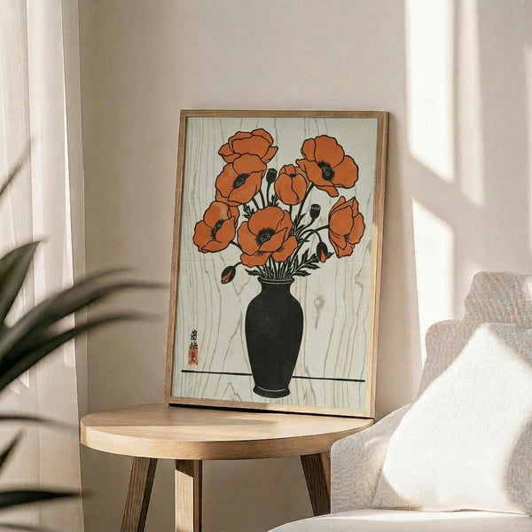 May include: Framed vintage-style botanical print featuring orange poppies in black vase displayed on round wooden side table next to beige upholstered chair. Artwork shows Japanese woodblock print aesthetic with orange flowers against light wood grain background. Natural lighting creates soft shadows on neutral wall. Plant leaves visible in corner.