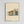 May include: Framed architectural poster showing modernist building illustration in muted green, orange, and cream colors against beige background. Features clean geometric lines, large windows, and flat roof with trees in background. Bauhaus text at bottom. Light wood frame mounted on white wall.