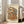 May include: Framed poster print leaning against white wall beside beige armchair in bright living room with large windows. Artwork depicts charming two-story Japanese ramen shop with warm orange and wood tones, traditional blue tile roof, outdoor seating with wooden stools, potted plants, and Japanese signage. Illustration has vintage watercolor style on cream background with power lines overhead. Light wooden frame complements neutral home decor.