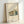 May include: Framed modernist architectural poster featuring a geometric mid-century house design in muted green, orange and blue tones against cream background. Natural wood frame leans against white wall with soft natural lighting creating diagonal shadows across the minimalist artwork.