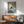 May include: Framed poster artwork showing a black cat reading a book while sitting on striped bedding in a cozy bedroom scene, displayed above a wooden sideboard with white ceramic lamp in a modern living room with sage green upholstered chair and round coffee table, natural sunlight casting geometric shadows on white wall