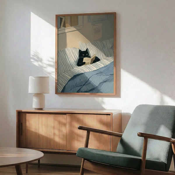 May include: Framed poster artwork showing a black cat reading a book while sitting on striped bedding in a cozy bedroom scene, displayed above a wooden sideboard with white ceramic lamp in a modern living room with sage green upholstered chair and round coffee table, natural sunlight casting geometric shadows on white wall