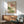 May include: Framed botanical print displaying pink poppies with green leaves and stems in vintage Japanese art style, hanging above a wooden sideboard with table lamp. Mid-century modern teal upholstered chair with wooden arms sits nearby on hardwood floor, with natural sunlight casting shadows across white wall.