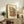 May include: Framed artwork displaying a charming two-story Japanese ramen shop with warm orange and blue tones, wooden details, hanging lanterns, and power lines above, shown in a cozy living room setting on a rustic wooden sideboard alongside books, a ceramic vase, and fern plant, with a beige upholstered chair visible in the foreground.