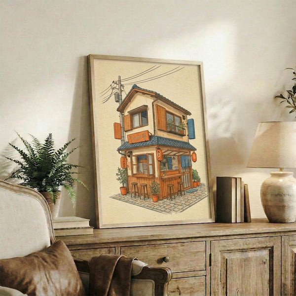 May include: Framed artwork displaying a charming two-story Japanese ramen shop with warm orange and blue tones, wooden details, hanging lanterns, and power lines above, shown in a cozy living room setting on a rustic wooden sideboard alongside books, a ceramic vase, and fern plant, with a beige upholstered chair visible in the foreground.