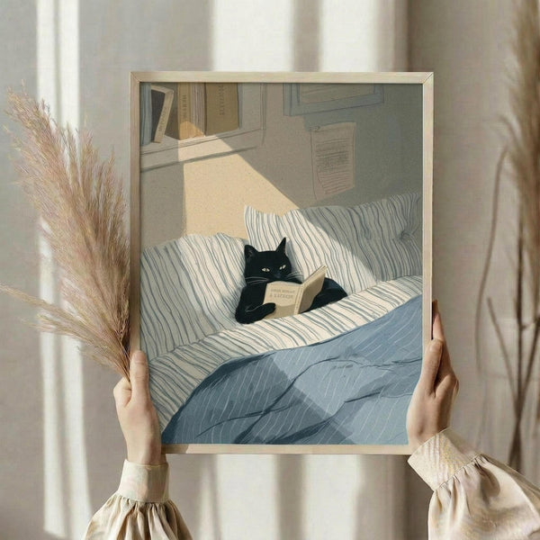 May include: Person holding framed artwork showing black cat reading book while lying on striped bedding. Scene features cozy bedroom with bookshelf, warm sunlight streaming across blue and white striped pillows and blanket. Dried pampas grass visible on left side. Artwork displayed in light wood frame against neutral bedroom setting.